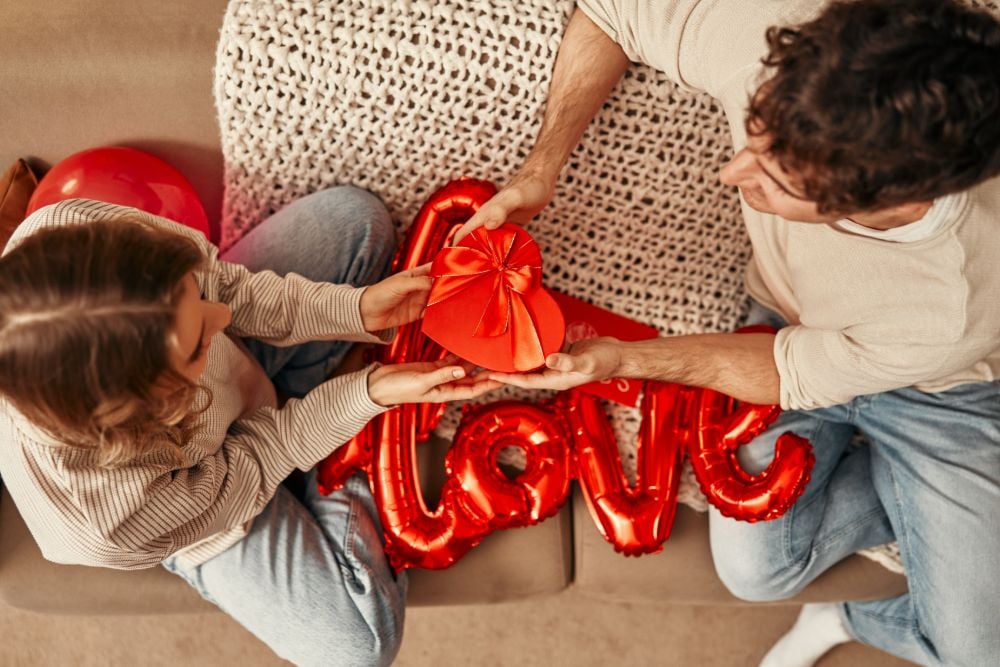 A top-down shot of a young couple sitting on a sofa with red balloons, one says "Love," and holding a heart-shaped red box with a bow.