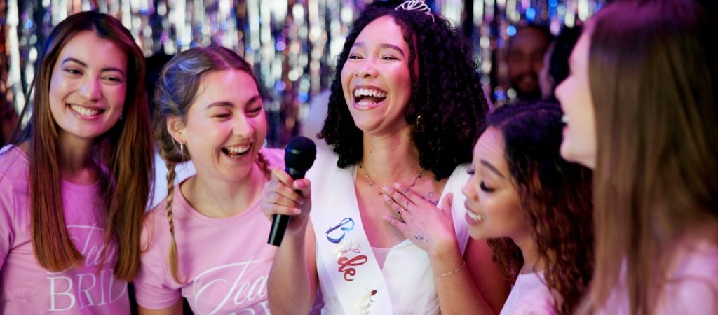 A joyful bride-to-be in a sash and tiara laughs with four friends gathered around a microphone singing karaoke.