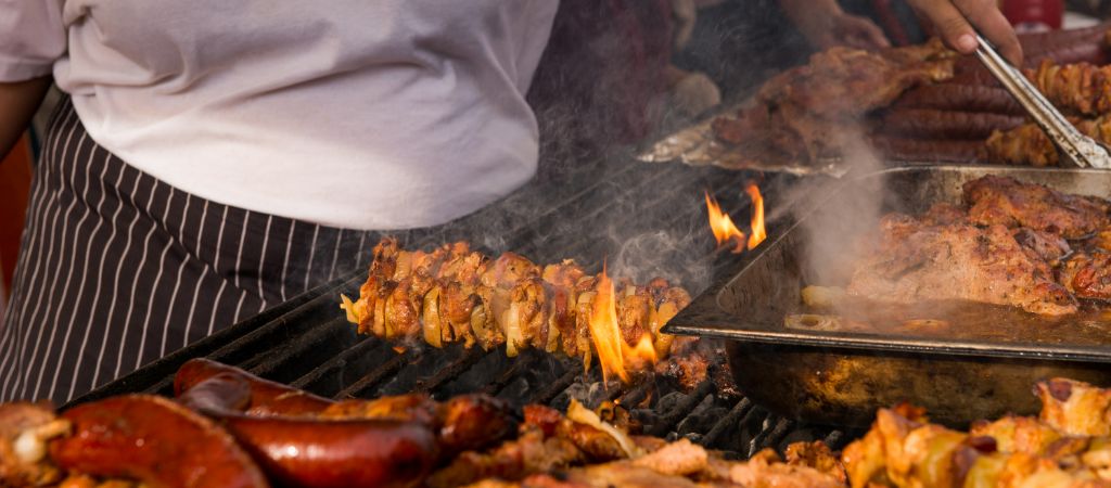 Person grilling meat skewers and various cuts of meat on an outdoor barbecue, with visible flames and smoke rising from the grill.