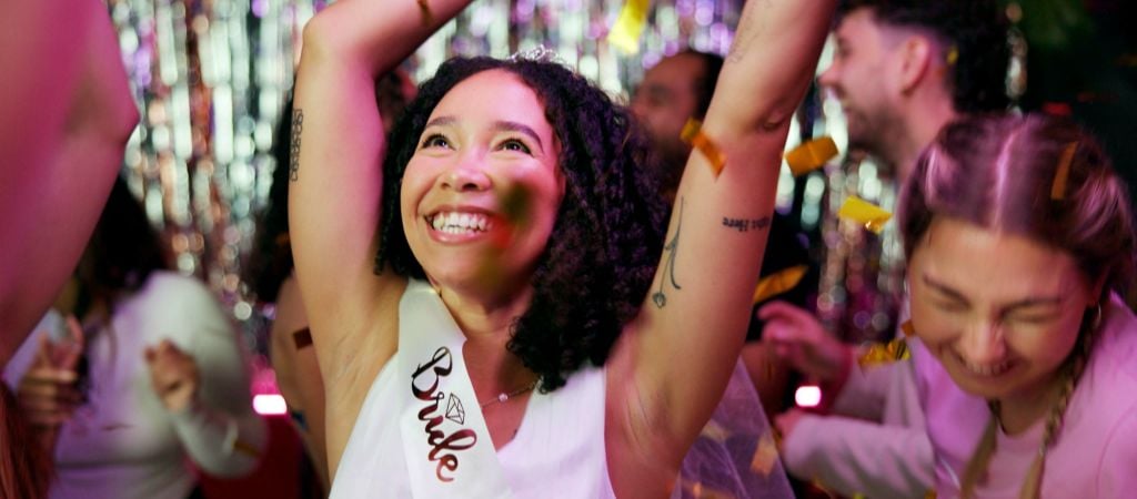 A joyful woman with a bride-to-be sash dances amid colorful confetti, surrounded by friends.