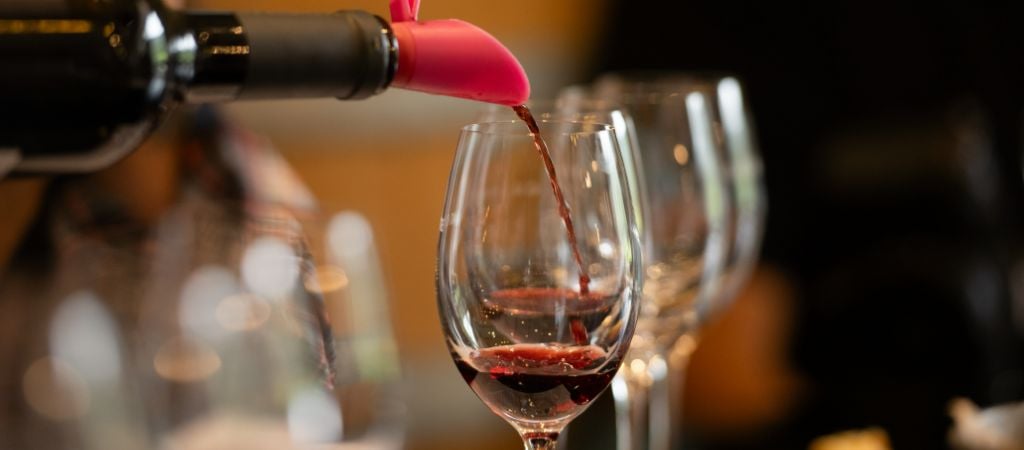 A bottle of red wine being poured into a row of stemmed glasses.