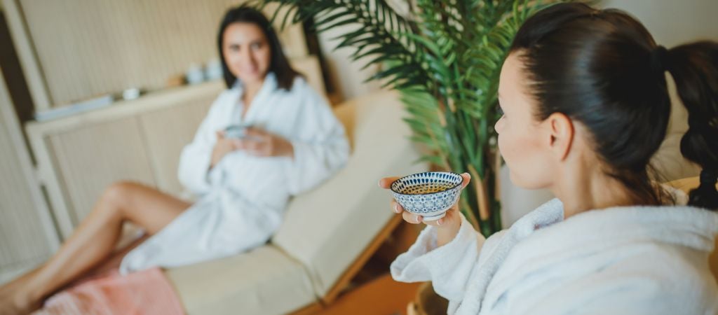 Two women relax in lounge chairs wearing white bathrobes in a spa.