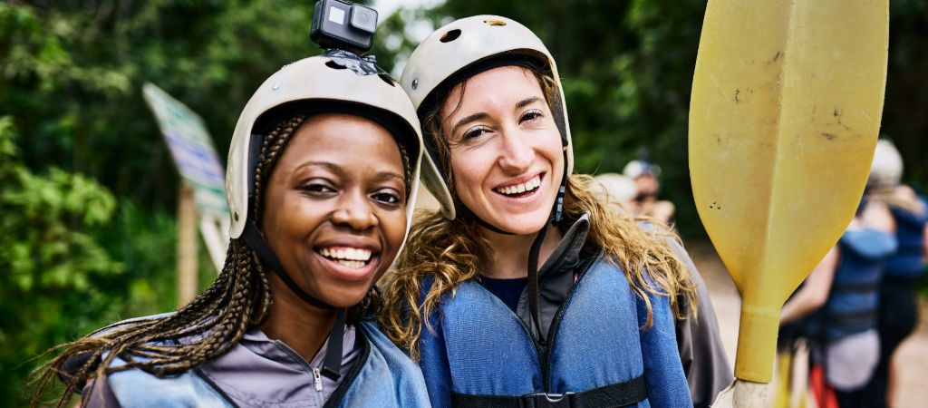 Two smiling young women dressed in whitewater rafting gear, wearing a GoPro and holding a paddle upright.