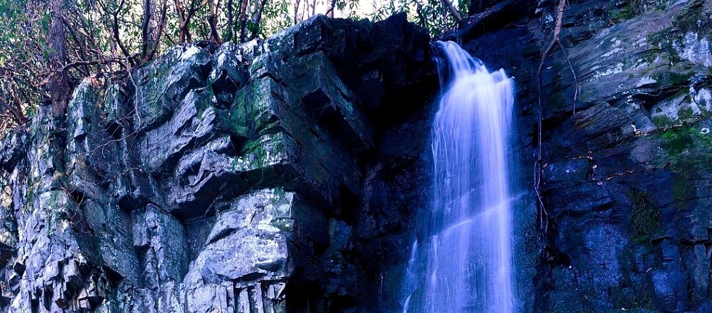 A graceful multi-tiered waterfall spills over mossy rock ledges into a rocky streambed deep in the forest of Great Smoky Mountains National Park.