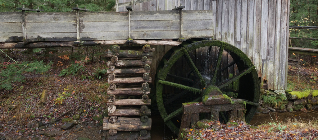 An old mill in Cades Cove moves against the water, with fall leaves scattered around the forest floor.