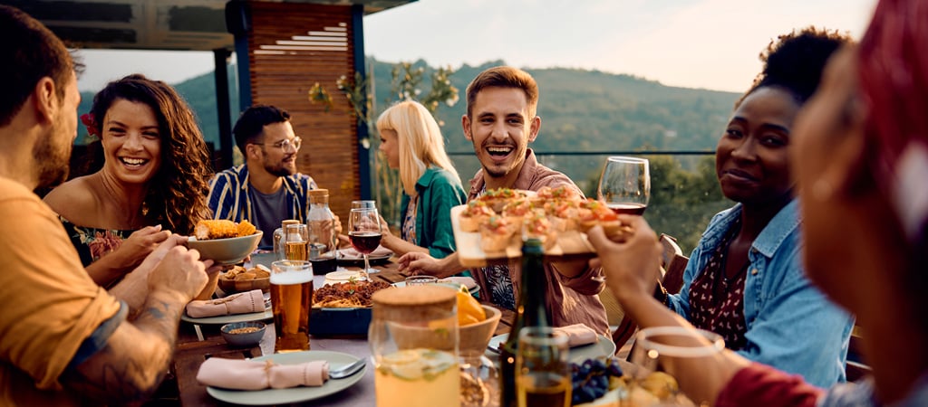 Cheerful Friends Enjoying a Meal Outside A group of friends laugh and smile while dining at an outdoor table, with mountain views in the background.