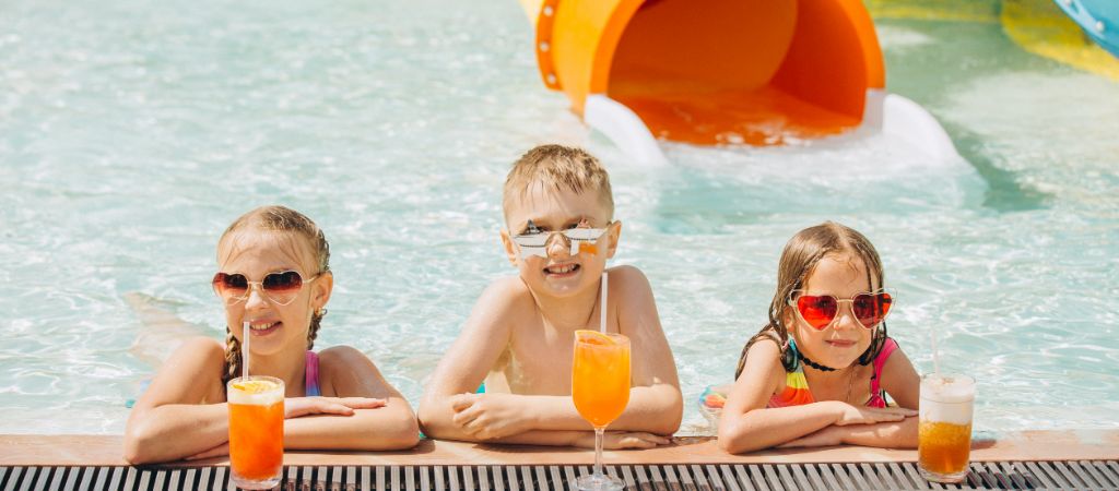 Three kids in a pool by drinks, smiling near a water slide.