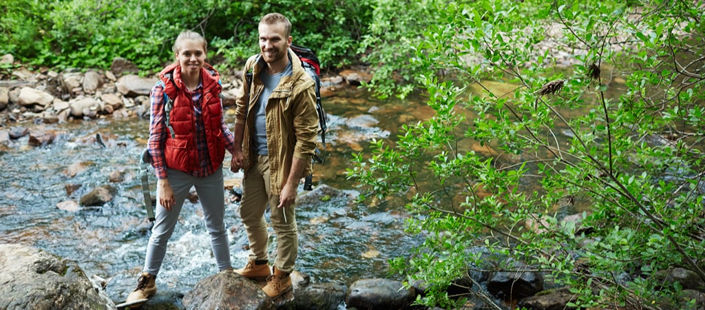 A young woman and man dressed in fall hiking gear hold hands while smiling at the camera and standing on stones in a creek.