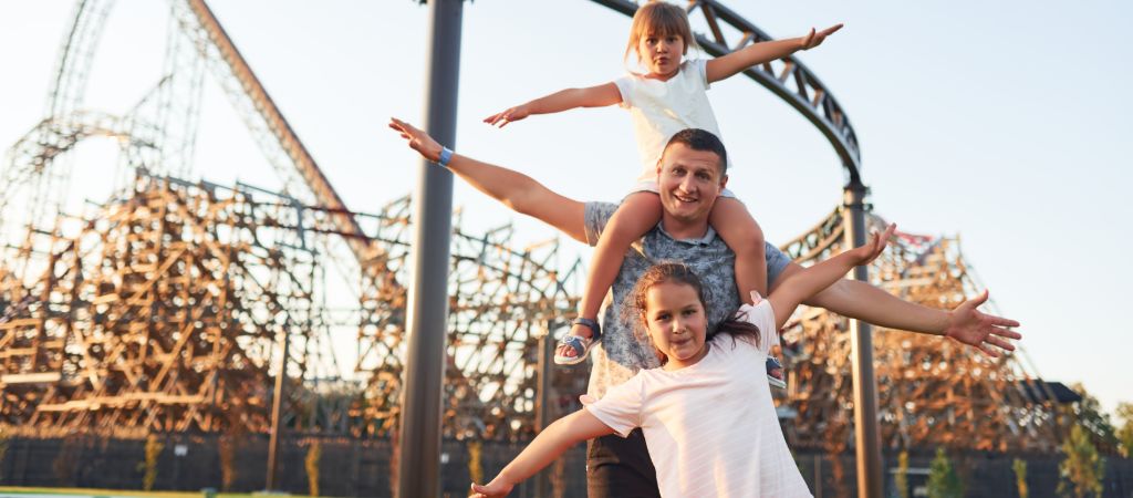 Father with two daughters posing playfully in front of a roller coaster.