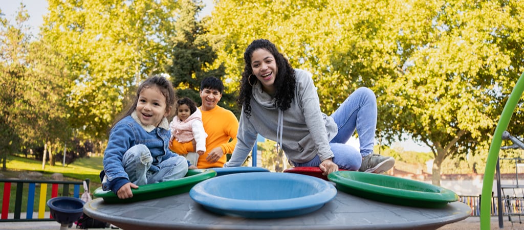 A young mother and daughter smiling while playing on a playset at a local playground on a sunny day, with the father and younger daughter smiling in the background.