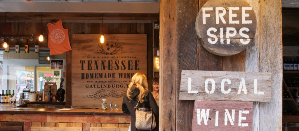 Free Wine Sips Sign in Downtown Gatlinburg A sign reads Free Sips, Local Wine at the Tennessee Homemade Wines shop in downtown Gatlinburg, with a female customer ordering a wine tasting in the background.