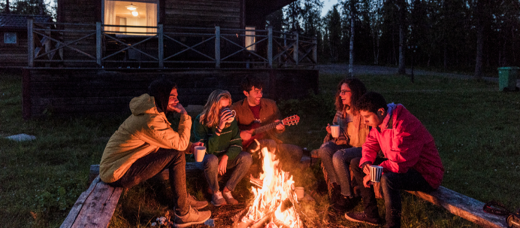 Friends Sitting Around a Campfire at a Cabin A group of young friends sit and sip around a campfire in front of a log cabin, with the center individual playing a guitar.