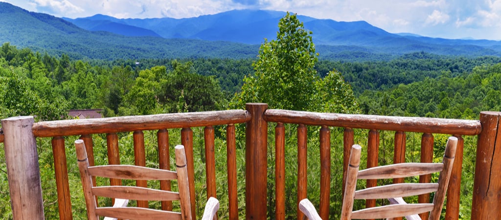 Two rocking chairs sitting on a Gatlinburg cabin deck face a panoramic view of the Great Smoky Mountains on a sunny day.