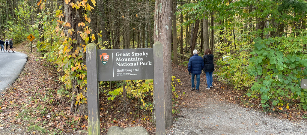 A Great Smoky Mountains National Park sign reads Great Smoky Mountains National Park, Gatlinburg Trail, National Park Service, U S Department of the Interior, with visitors hiking nearby.