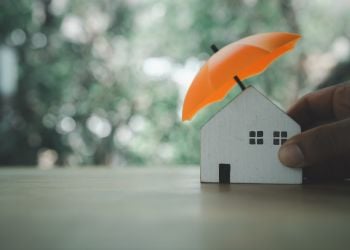 Man's hand holds an orange umbrella over a white wooden block shaped like a house, with blurred trees in the background.