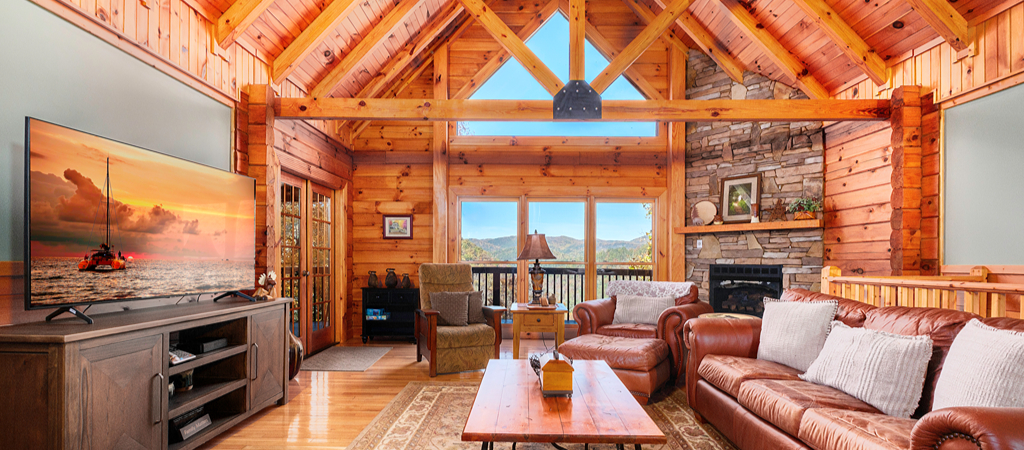 Pigeon Forge Cabin Living Room An interior of a Pigeon Forge cabin's living room, with a big screen TV, sofa, stacked-stone fireplace, and windows overlooking a Smoky Mountain view on a sunny day.