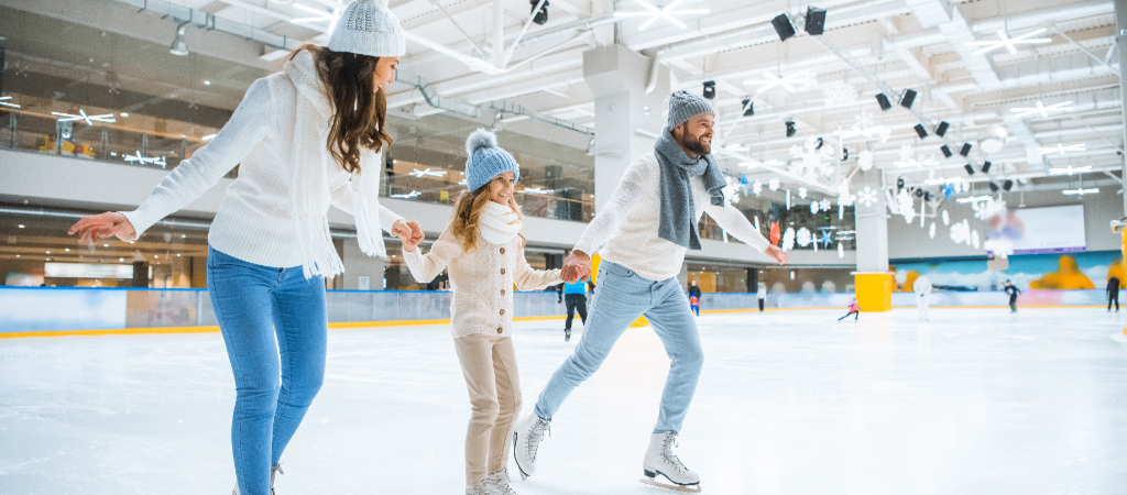 Smiling Family Holding Hands While Ice Skating A young mother, father, and daughter dressed in winter wear smile while ice skating at an indoor rink.