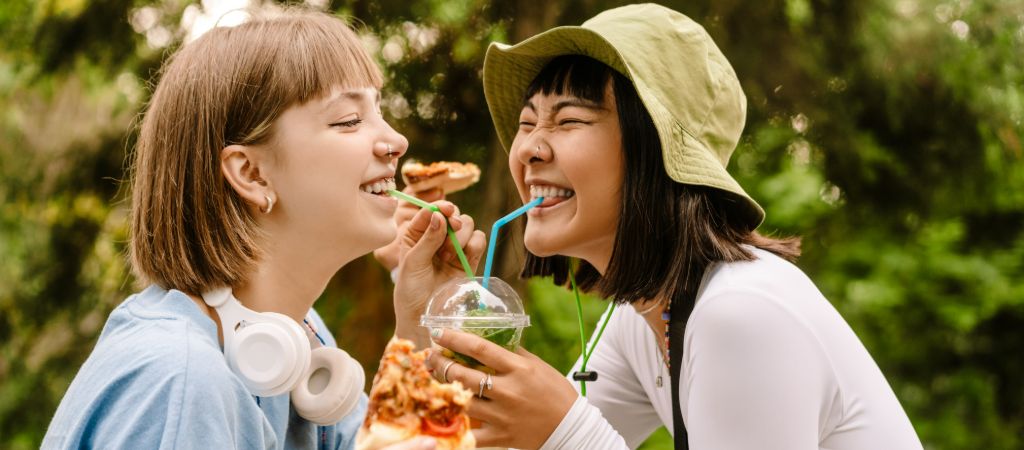 Two smiling girls sharing a drink and food outdoors.