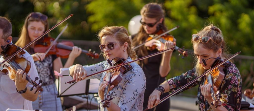 Group of violinists performing outdoors in sunlight.