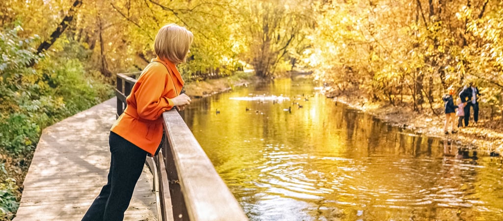 A woman on a wooden path leans over a barrier to peer at the beautiful river reflecting fall colors.